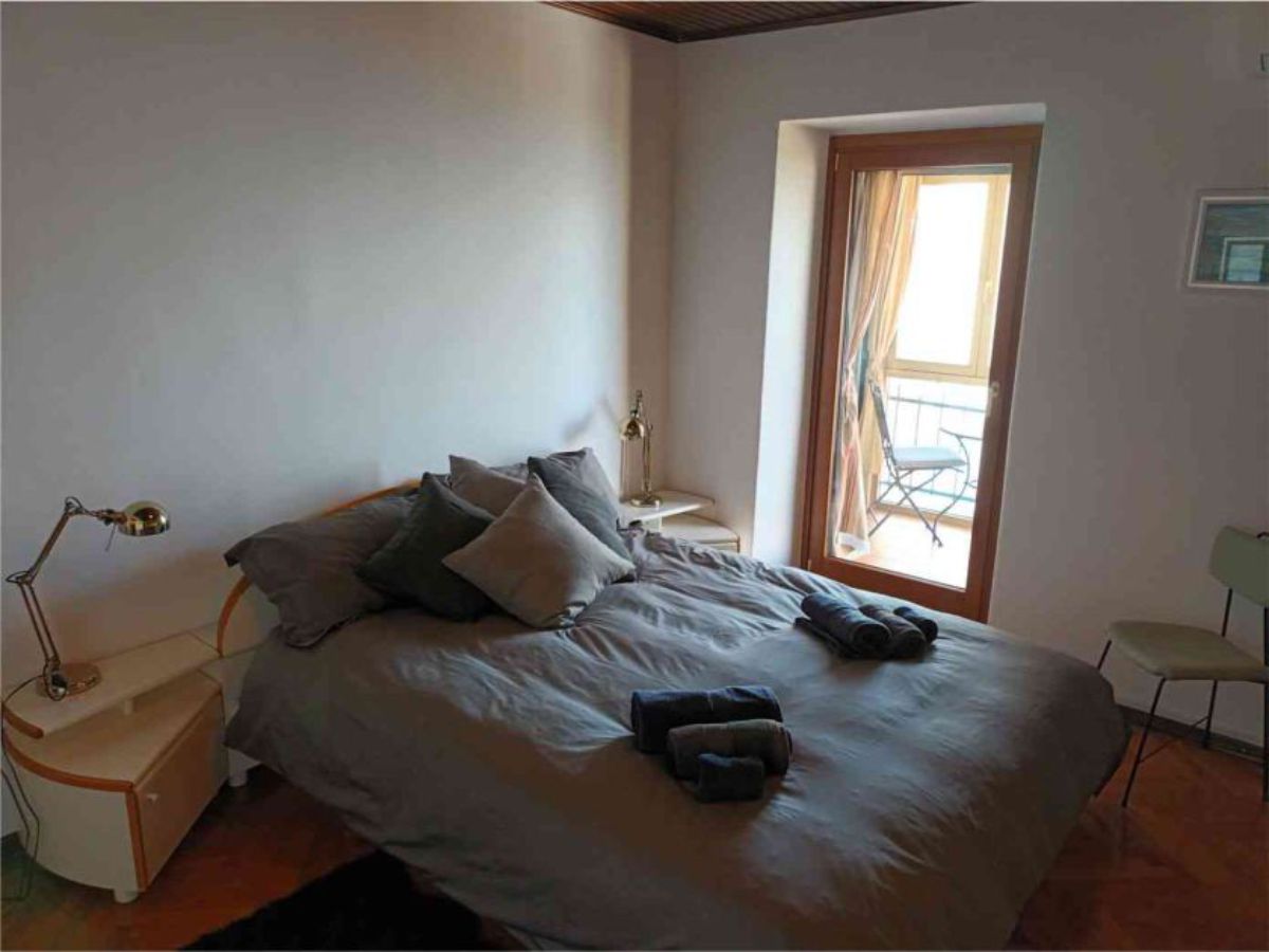 room with a double bed with pillows and a bedspread, four rolled-up towels, two bedside tables with lamps, a door in the background 