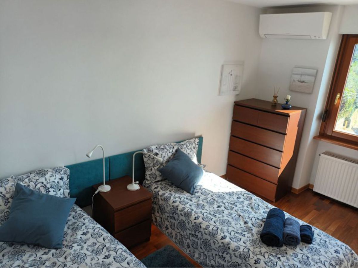room with two separate beds with pillows, rolled towels, a bedside table with two lamps, a chest of drawers, wall-mounted air conditioning, a window
