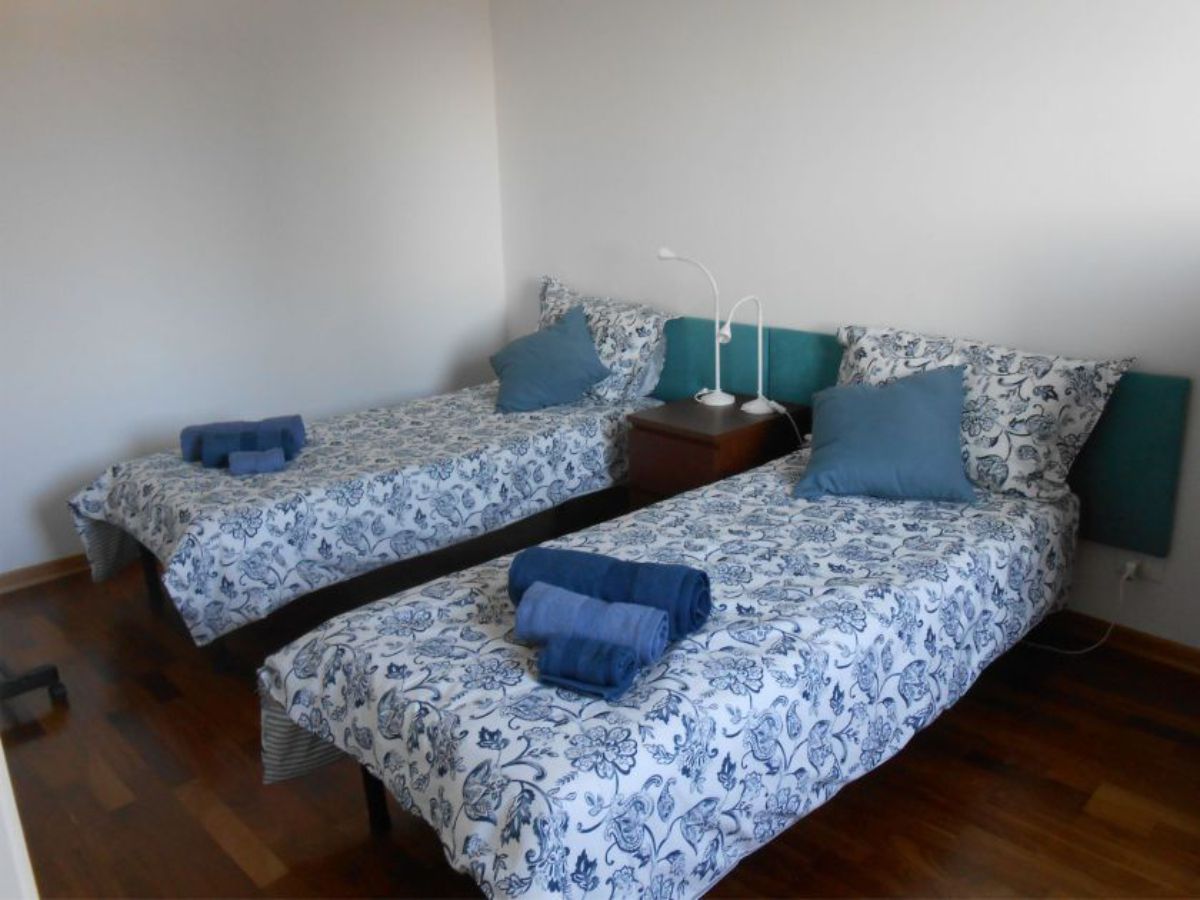 two beds with a bedspread and rolled up towels, pillows, a bedside table with lights in the middle