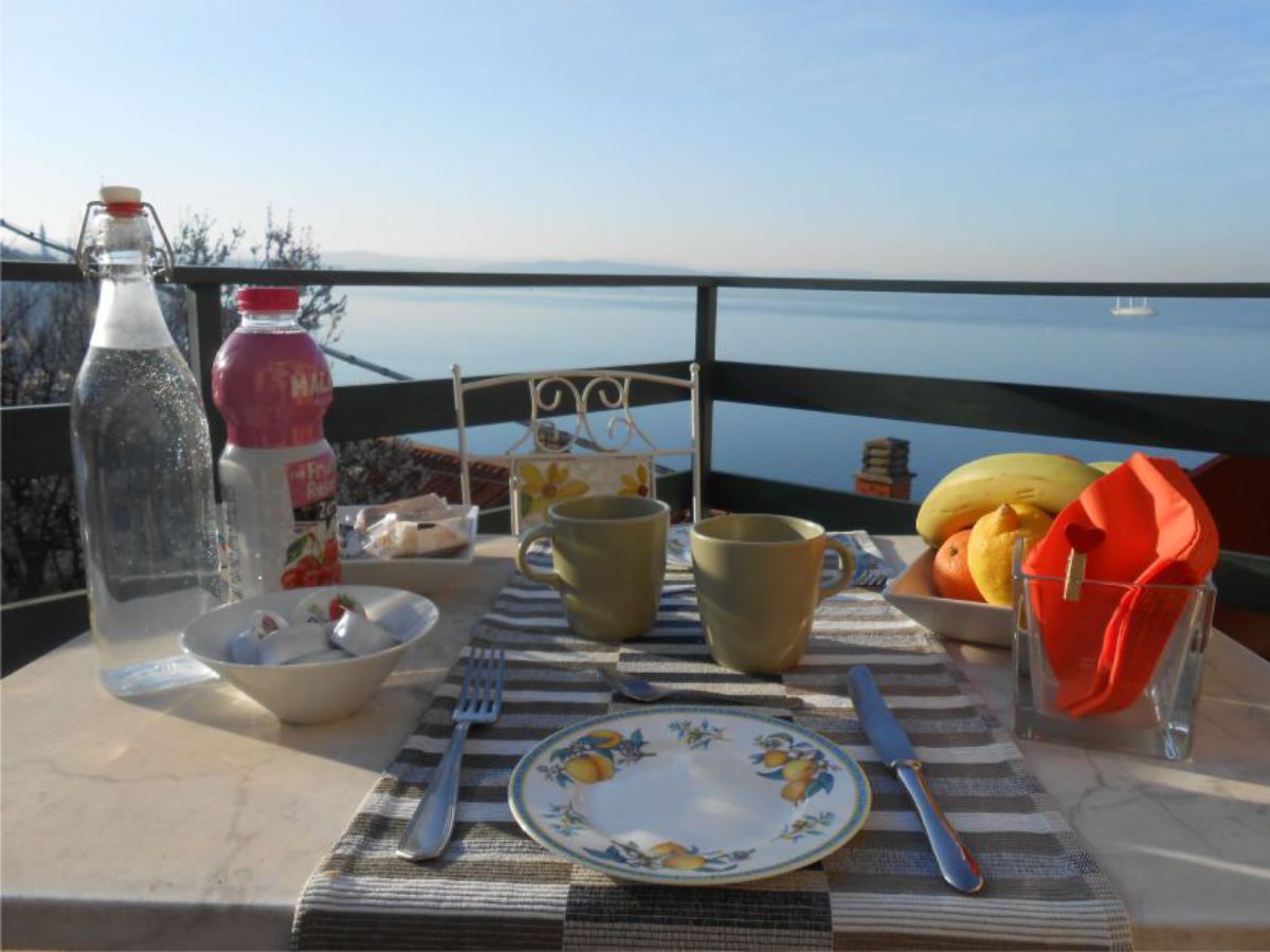 Table with plate, glasses, bottles, fruit, balcony railing, sea in the background